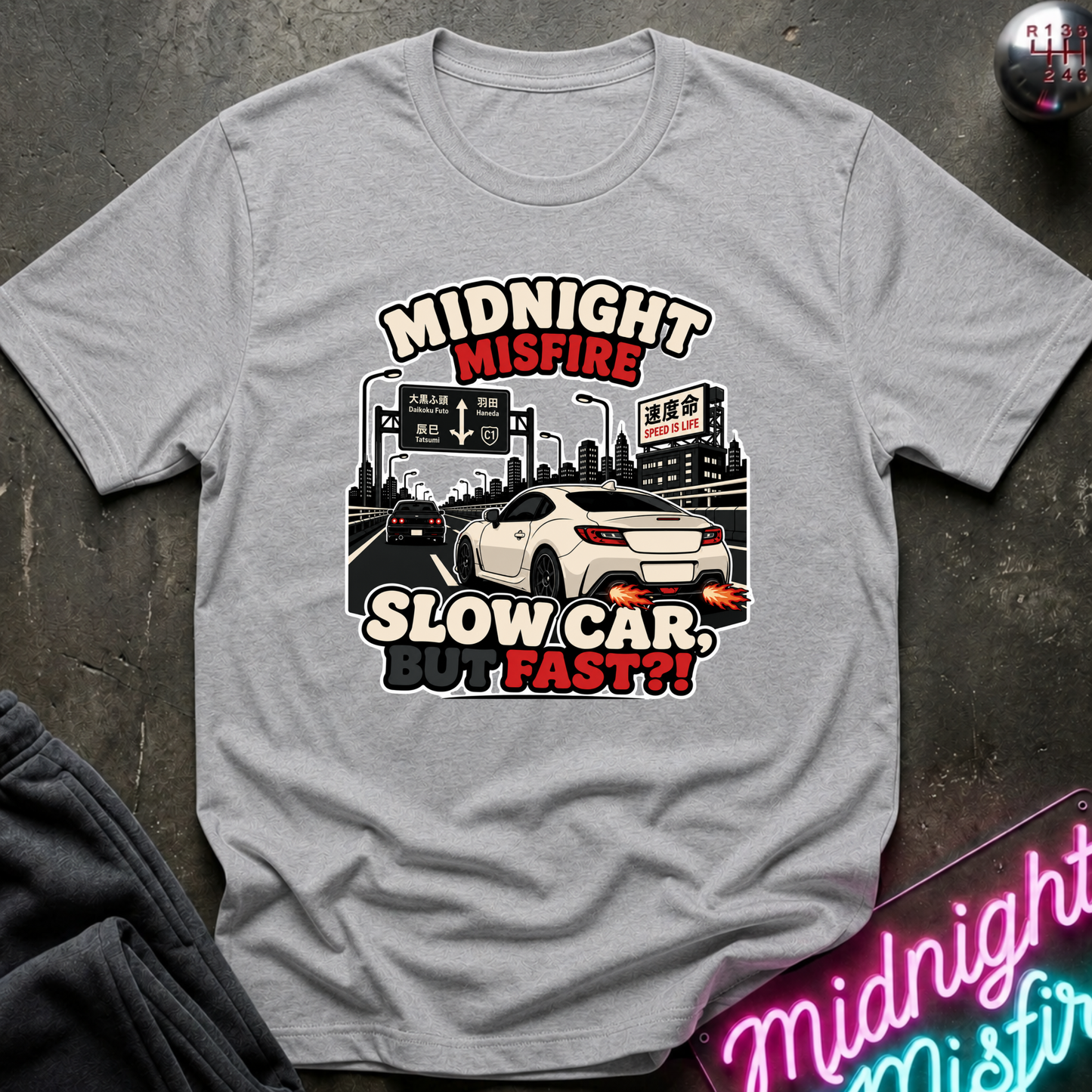 A light gray crewneck t-shirt featuring a graphic print of a white sports car on a highway with the text 'MIDNIGHT MISFIRE SLOW CAR BUMP FAST??'. The shirt is displayed flat on a dark textured surface with a neon sign and dark pants partially visible as props.