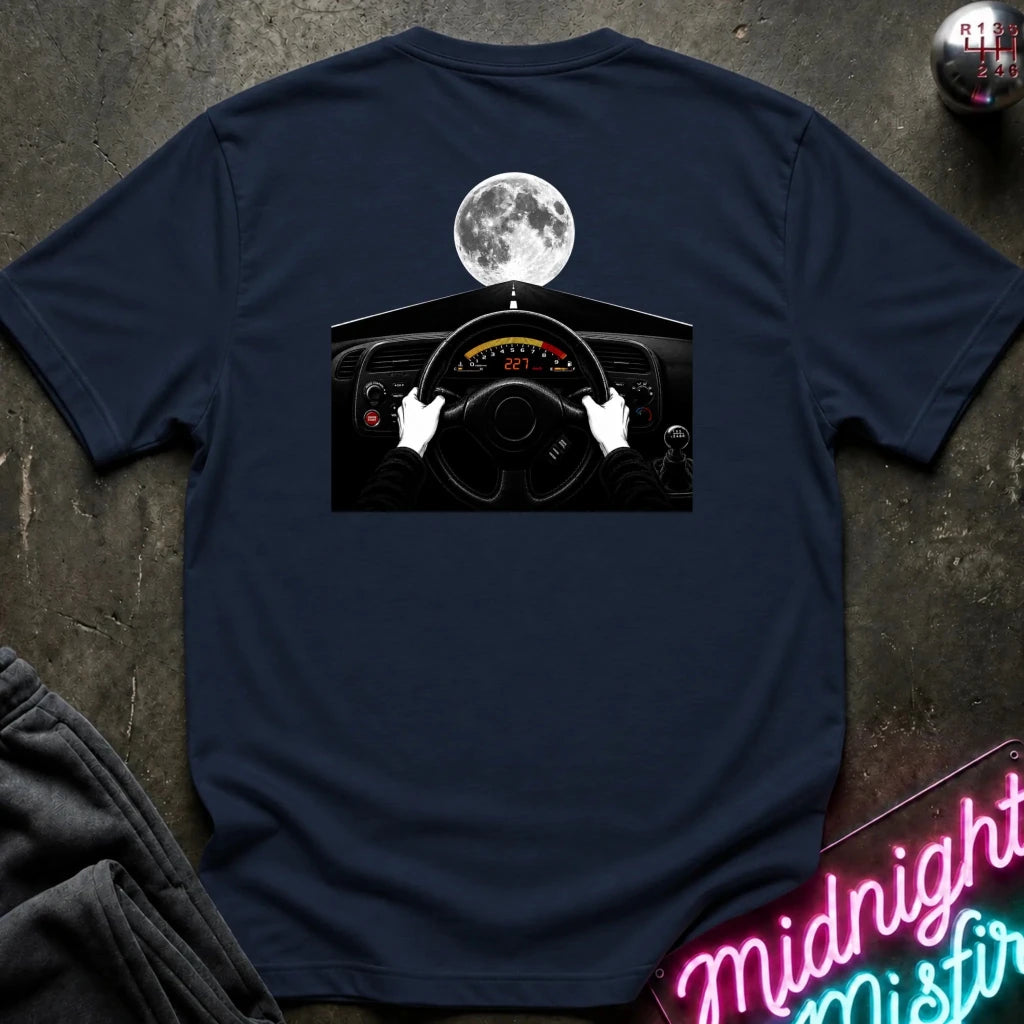 A navy blue short-sleeve t-shirt laid flat on a dark textured surface, featuring a graphic print of hands on a car steering wheel with a full moon above. 