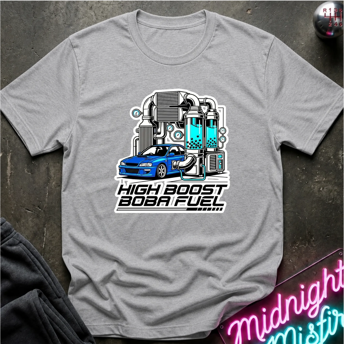 A heather gray short-sleeve t-shirt laid flat on a dark textured surface, featuring a graphic of a blue car connected to a high-tech lab setup with the text 'HIGH BOOST BOBA FUEL'.