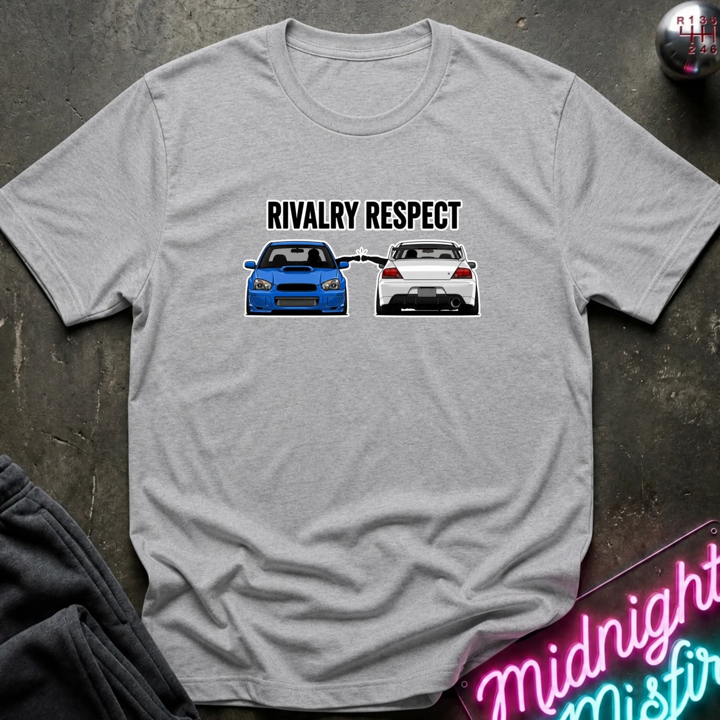 A light gray heather short-sleeve graphic t-shirt laid flat on a dark textured surface. The graphic features the text 'RIVALRY RESPECT' above a blue and white car, 