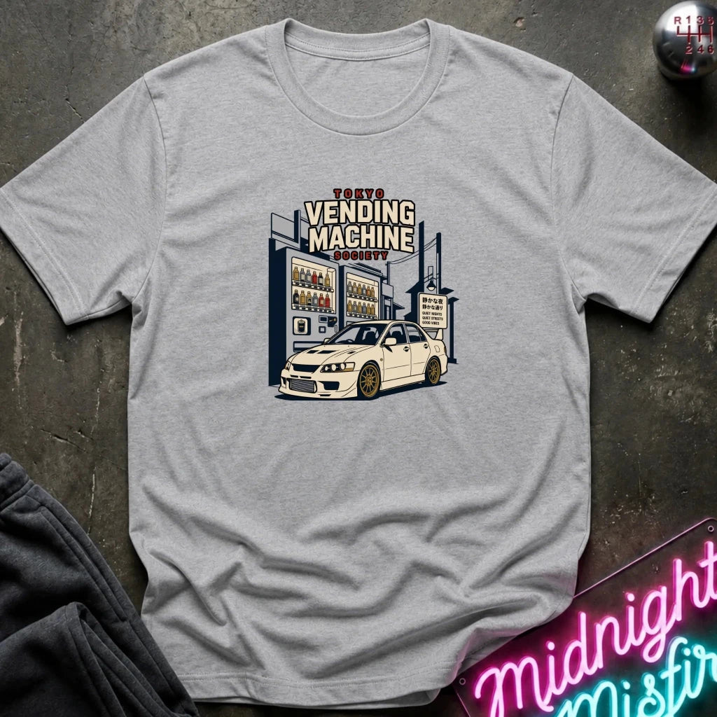 A light gray heather crewneck t-shirt featuring a retro-style graphic of a white sports car parked in front of a vending machine labeled 'Tokyo Vending Machine Society'. The shirt is laid flat on a dark textured surface with a neon sign and dark pants partially visible in the corners.