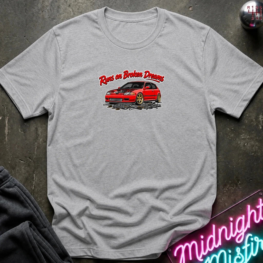 A light gray heather crewneck t-shirt laid flat on a dark textured surface. The shirt features a graphic print of a red sports car with the text 'Runs on Broken Dreams' in red stylized lettering.