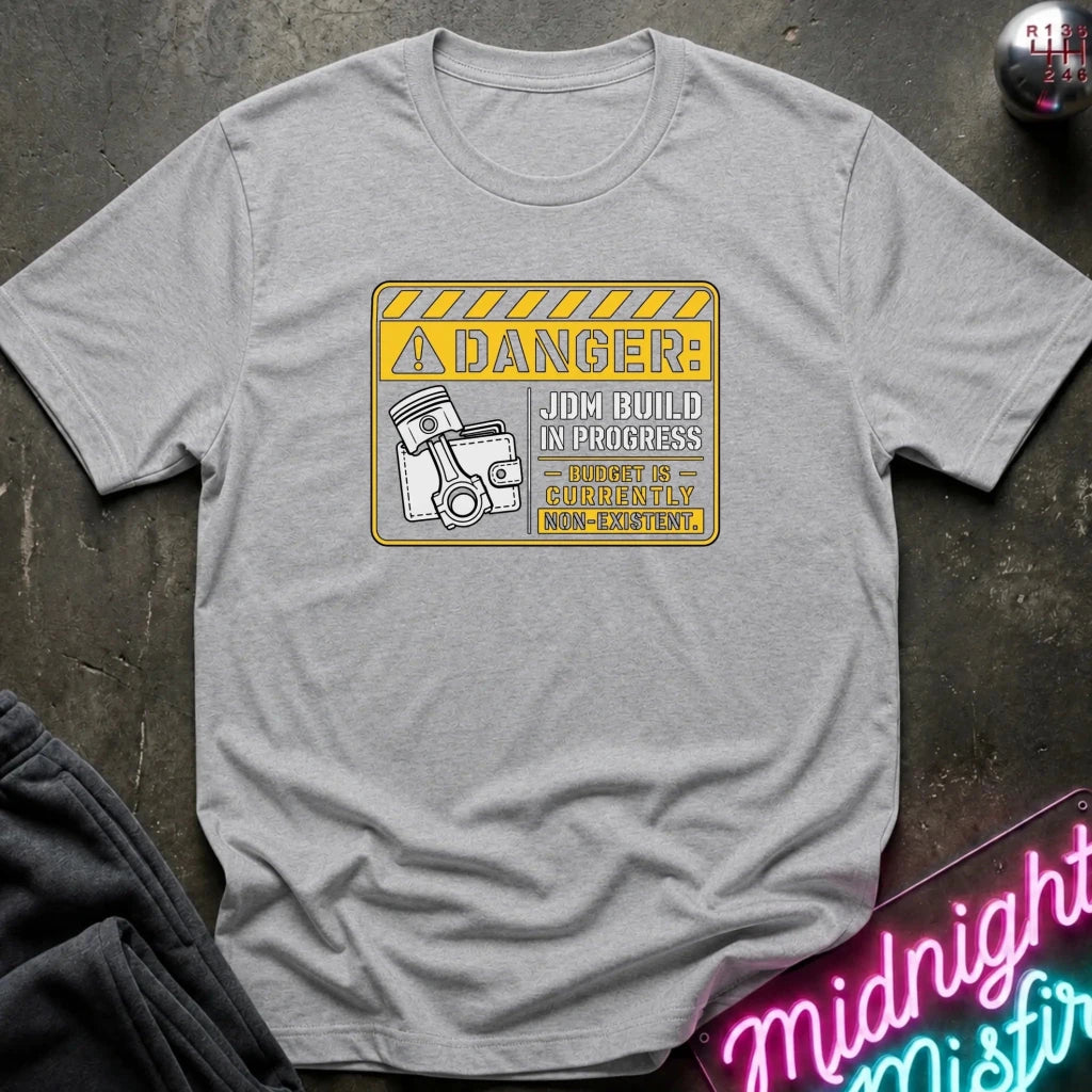 A grey short-sleeve crewneck t-shirt laid flat on a dark textured surface. The shirt features a yellow and black warning sign graphic on the chest with the text 'DANGER: JOB BUILD IN PROGRESS - 