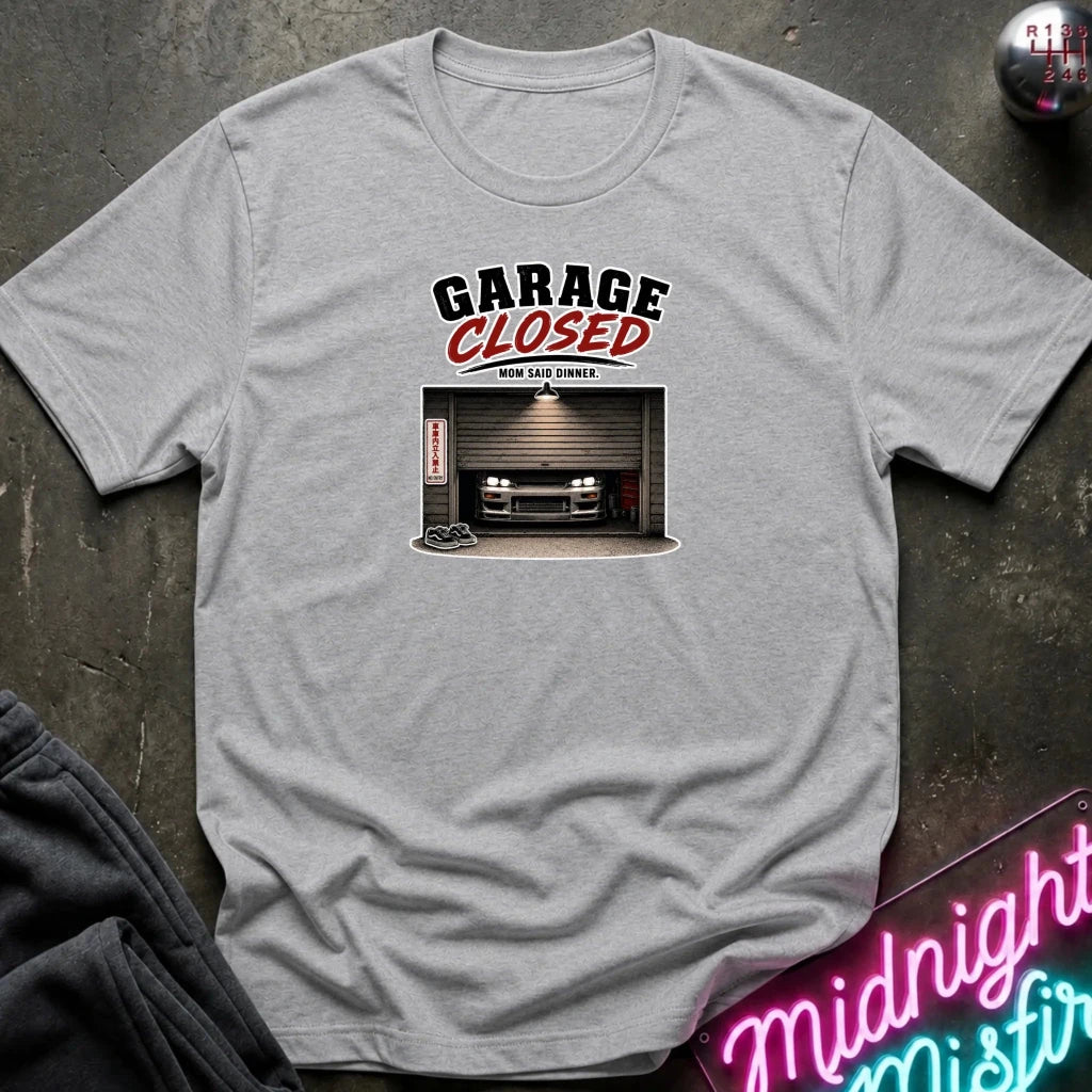 A light heather gray t-shirt laid flat on a dark textured surface, featuring a graphic print with the text 'GARAGE CLOSED' and a photo of a car parked inside a garage. 