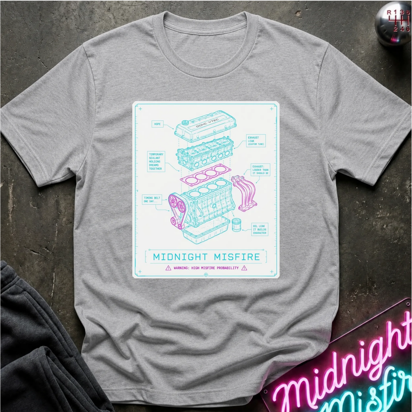 A heather gray short-sleeve t-shirt laid flat on a dark textured surface, featuring a central white graphic print with a light blue and pink technical-style engine diagram labeled 'MIDNIGHT MISFIRE.' A neon sign and folded dark fabric are partially visible around the edges as styling props.