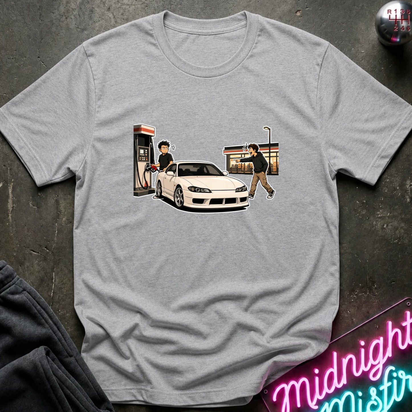 A light gray heather crewneck t-shirt featuring a graphic print of a white sports car at a gas station with two characters, styled flat on a dark textured background with a neon sign and dark pants partially visible.