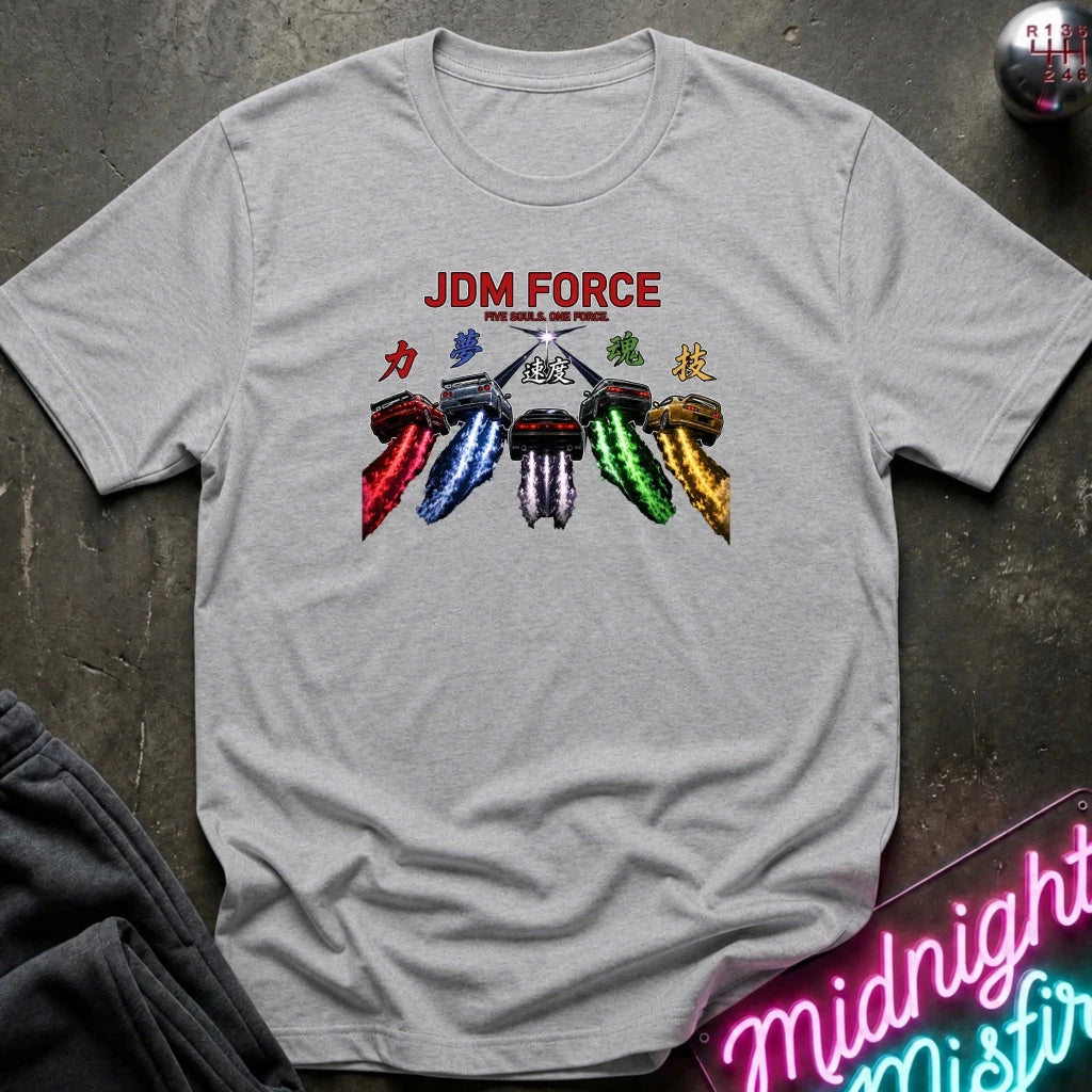 A light gray heather crewneck t-shirt featuring a graphic of six colorful motorcycle exhaust pipes with Japanese kanji symbols above the red text 'JDM FORCE'. The shirt is laid flat on a dark textured surface with a neon sign and dark pants partially visible in the corners.