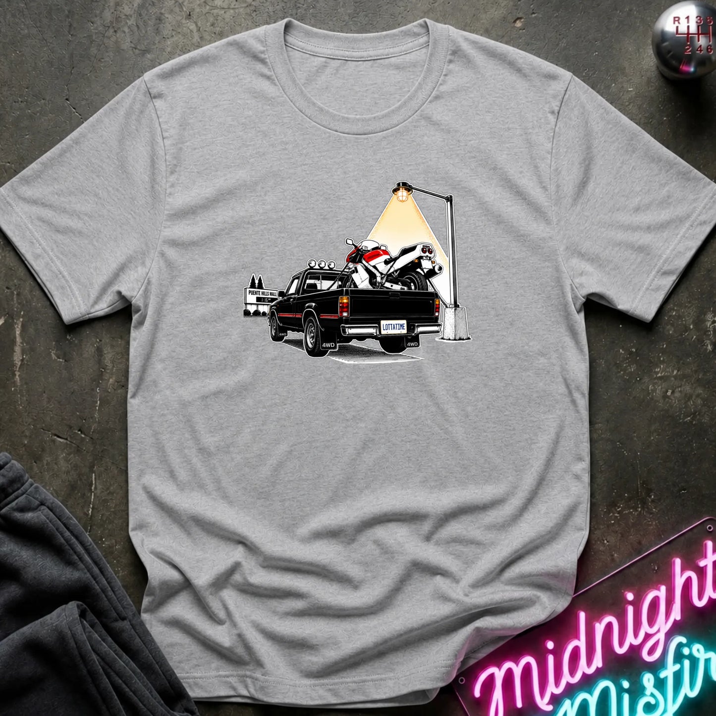 A heather gray short-sleeve crewneck t-shirt featuring a graphic print of a black pickup truck carrying a white and red motorcycle under a street lamp, laid flat on a dark textured surface.