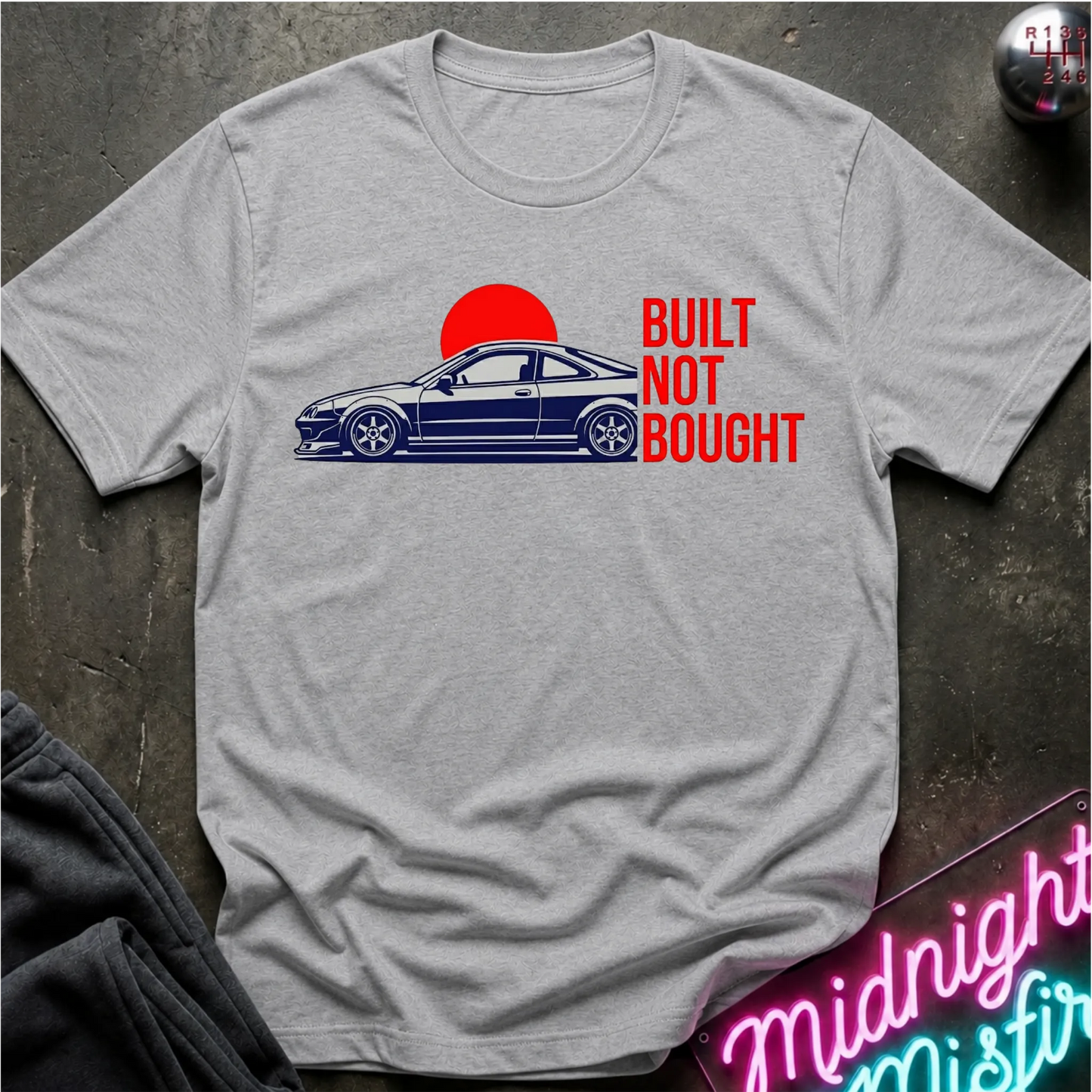 A light gray crewneck t-shirt laid flat on a dark textured surface. The shirt features a graphic print of a blue sports coupe car in front of a red sun, with the text 'BUILT NOT BOUGHT' printed in red.