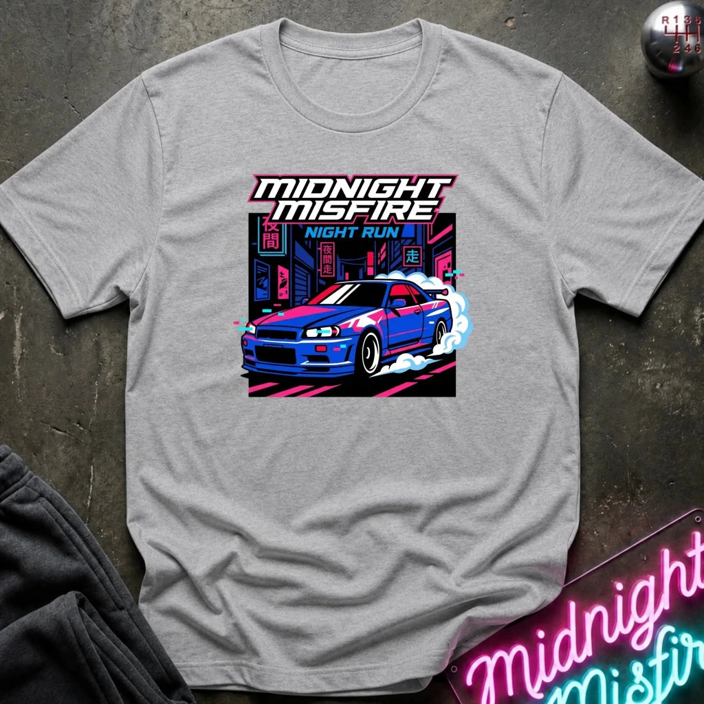 A light gray heather crewneck t-shirt featuring a colorful graphic print of a blue and red sports car with neon city lights and the text 'MIDNIGHT MISFIRE NIGHT RUN '. The shirt is laid flat on a dark textured surface with a neon sign and dark sweatpants partially visible.