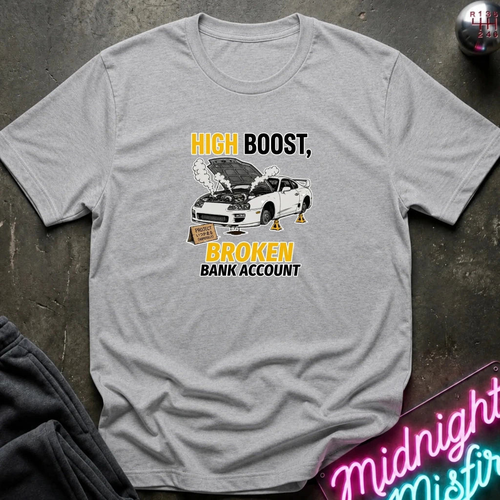 A light gray heather crewneck t-shirt with a graphic print of a white car with its hood open, emitting smoke, with the text 'HIGH BOOST, BROKEN BANK ACCOUNT' in yellow and black. The shirt is laid flat on a dark textured surface with a neon sign and dark pants partially visible.