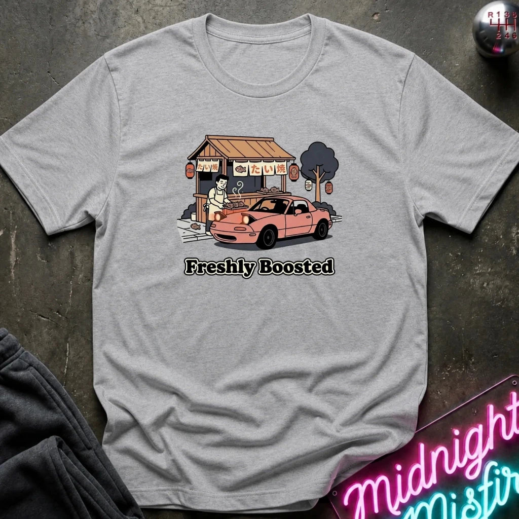 A heather gray short-sleeve t-shirt laid flat on a dark textured surface, featuring a graphic of a pink sports car parked in front of a small food stall, with the text 'Freshly Boosted' printed below.