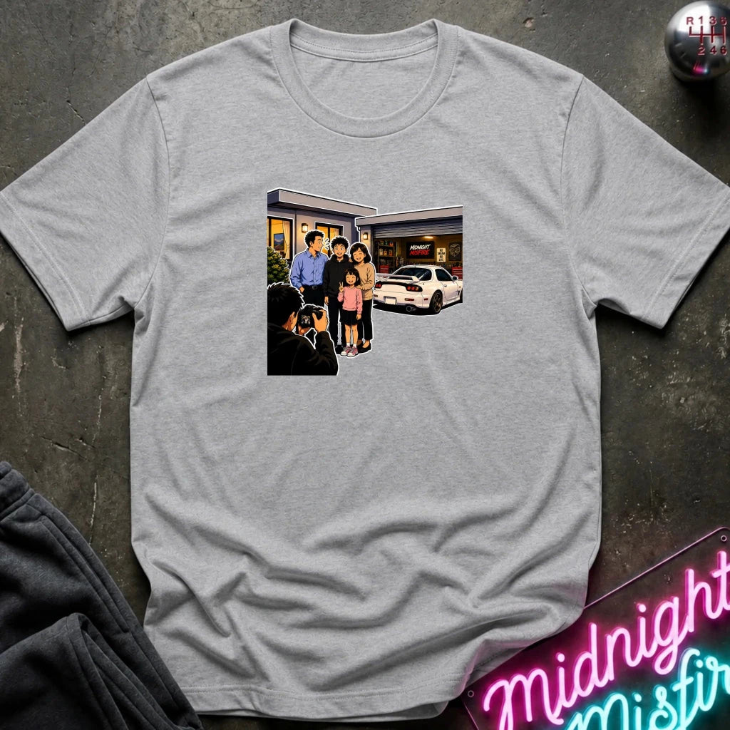 A light gray short-sleeve crewneck t-shirt laid flat on a dark textured surface, featuring a rectangular graphic print of a family standing outside a building taking a photo.