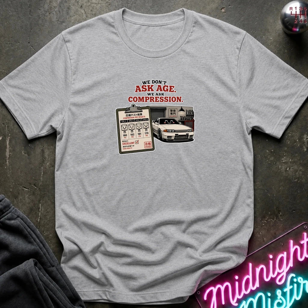 A light gray heather short-sleeve graphic t-shirt laid flat on a dark textured surface. The graphic features the text 'WE DON'T ASK AGE, WE ASK COMPRESSION.