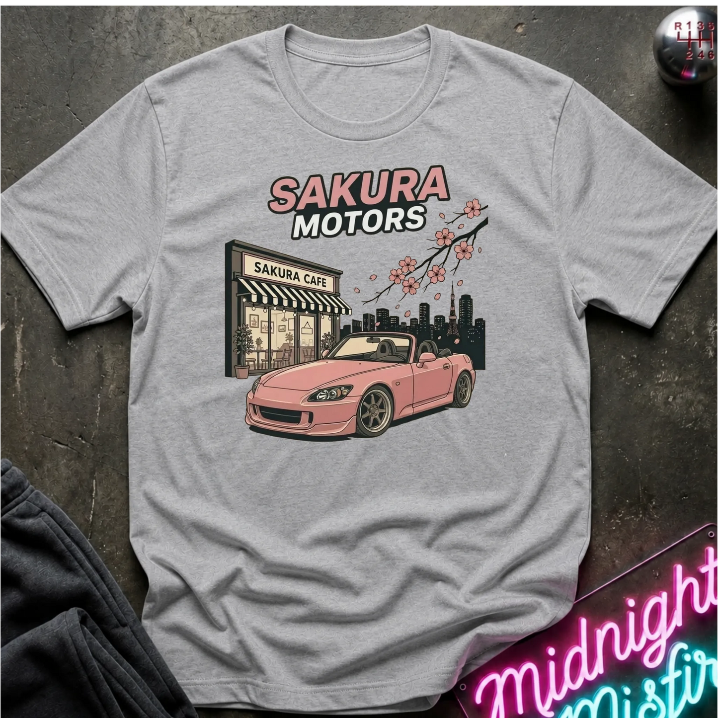 A light gray crewneck t-shirt laid flat on a dark textured surface, featuring a graphic print of a pink convertible sports car in front of a 'Sakura Motors' cafe and city skyline with cherry blossoms.