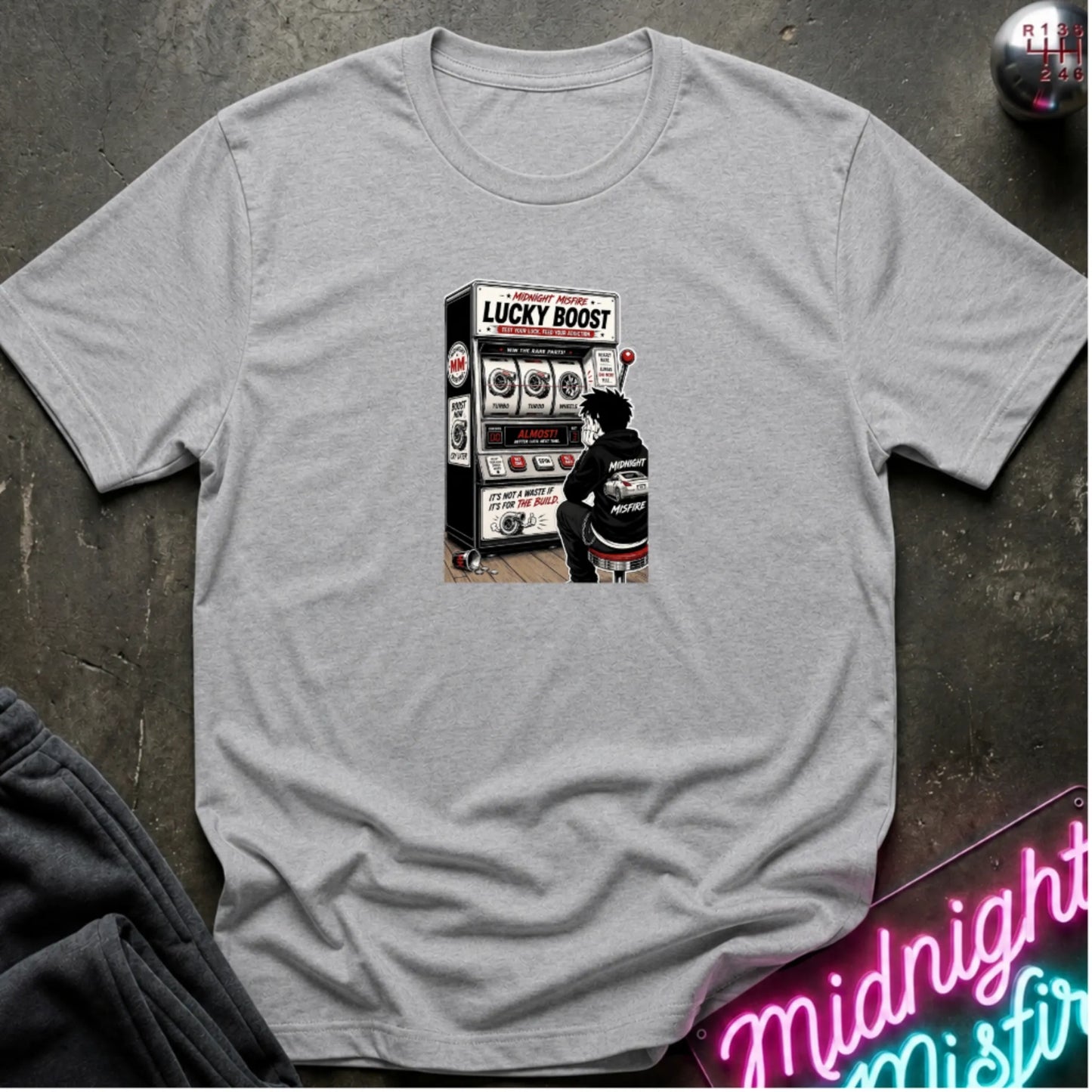 A heather gray  short-sleeve t-shirt with a retro-style graphic of a slot machine, displayed flat on a dark textured surface with a neon sign and sweatpants partially visible.