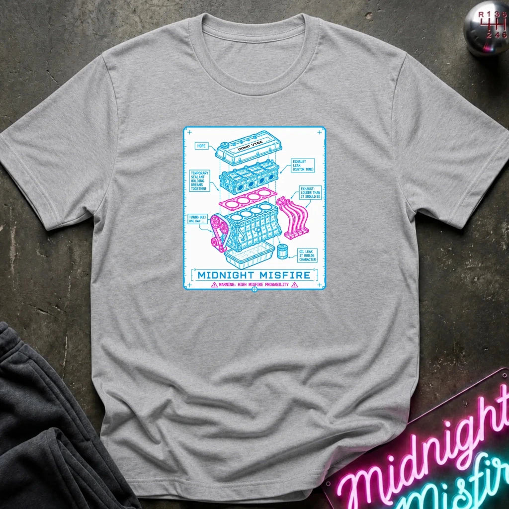gray short-sleeve graphic T-shirt laid flat on a dark textured surface. The shirt features a centered rectangular print with a light blue and pink exploded-style engine diagram and the text 'MIDNIGHT MISFIRE.