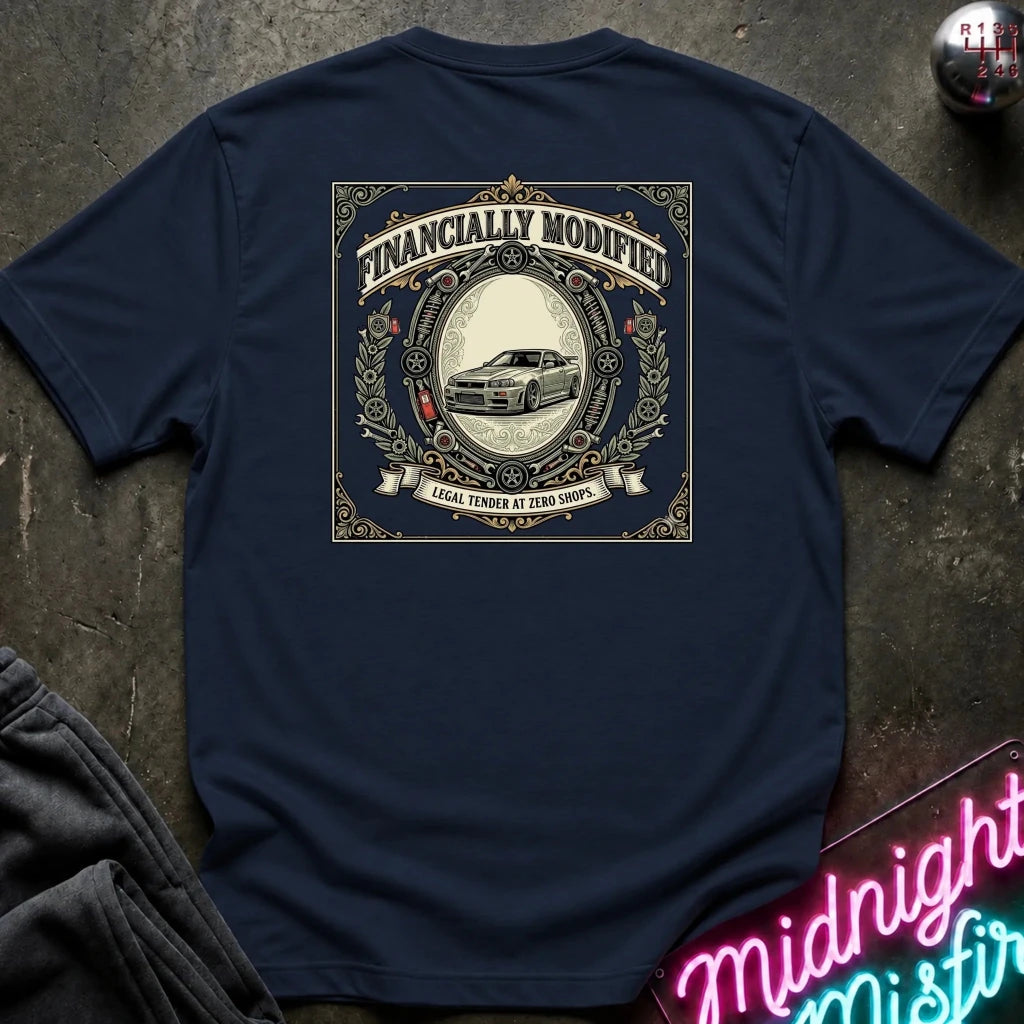 A navy blue t-shirt laid flat on a dark textured surface, showing the back graphic with the text 'FINANCALLY MODIFIED' and a vintage-style car illustration. A neon sign reading 'Midnight Mistir' is partially visible in the bottom right corner.