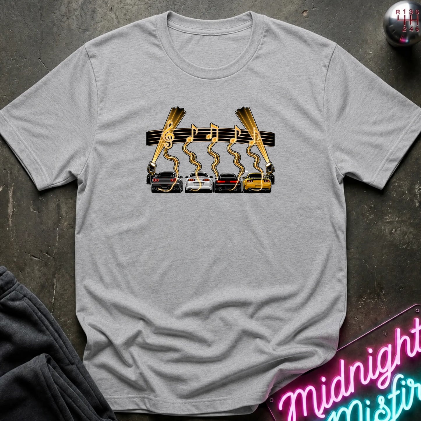 A light gray heather short-sleeve t-shirt laid flat on a dark textured surface, featuring a graphic print of four cars (red, white, gray, yellow) with gold wavy lines and musical notes above them, flanked by two  lights.