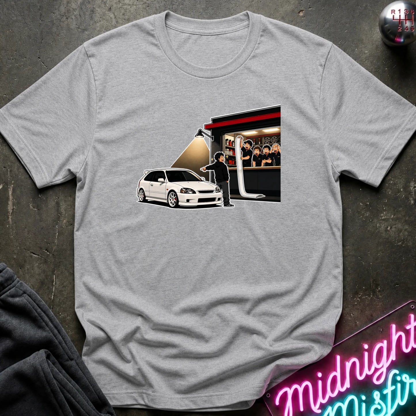 A light gray crewneck t-shirt laid flat on a dark textured surface, featuring a graphic print of a white hatchback car. A neon sign reading 'Midnight Misfire' is partially visible in the bottom right corner.
