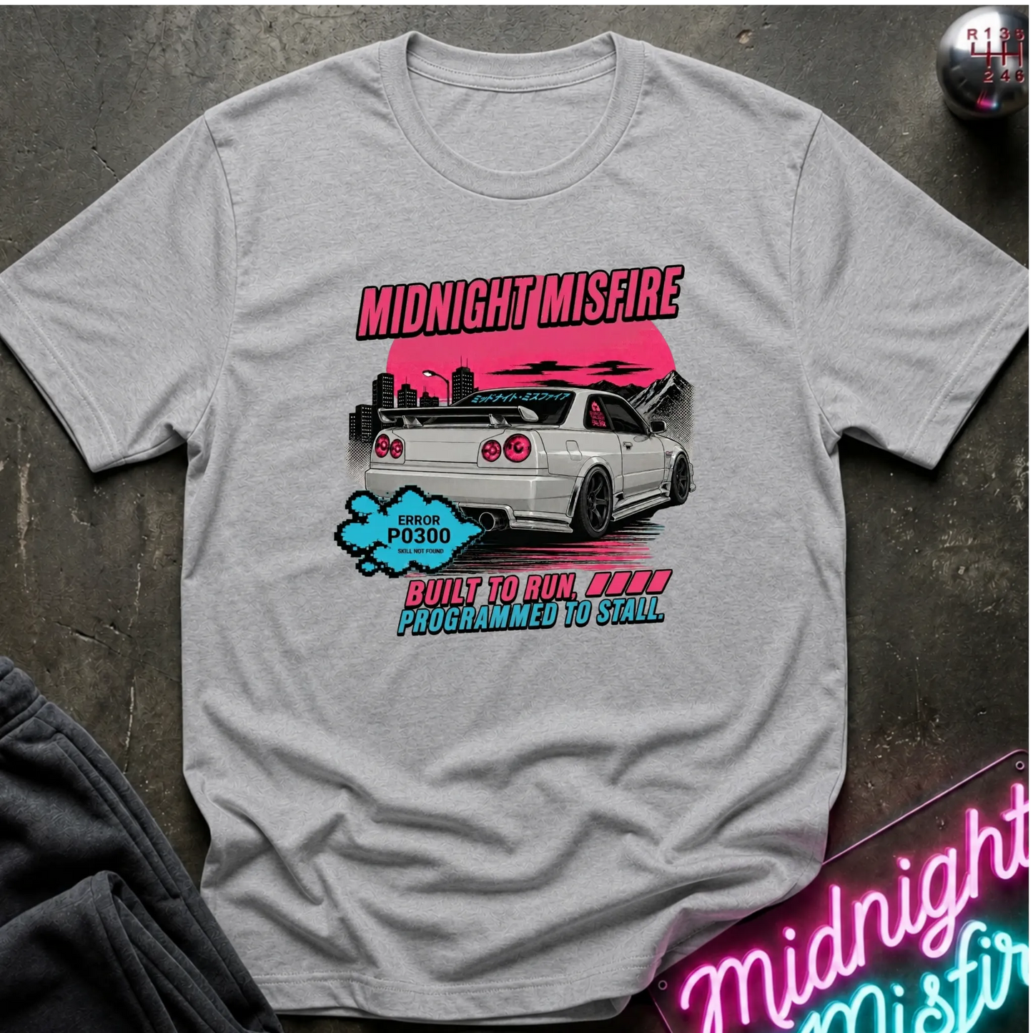 A light gray short-sleeve graphic t-shirt laid flat on a dark textured surface. The graphic features a white sports car with a pink sunset, the text 'MIDNIGHT MISFIRE' in pink.
