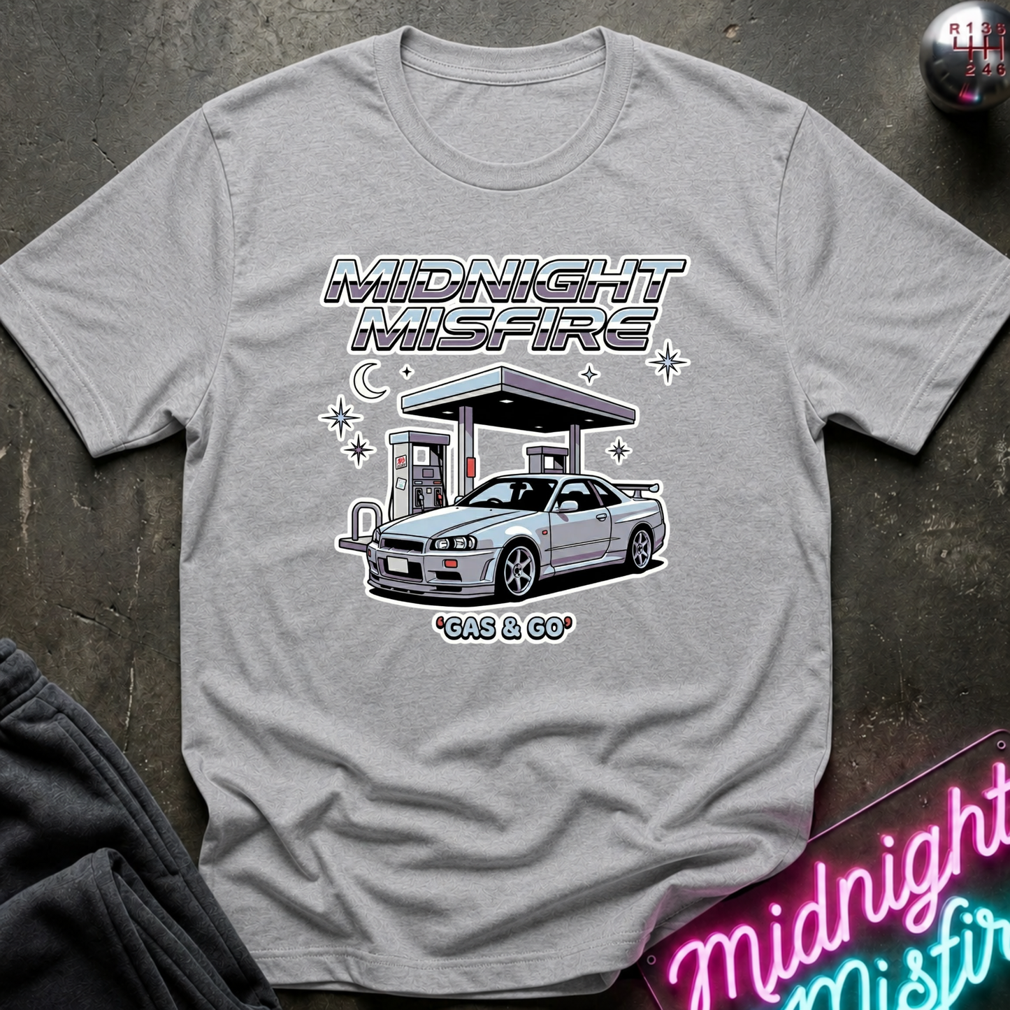 A light gray heather crewneck t-shirt featuring a graphic print of a silver sports car at a gas station under the text 'MIDNIGHT MISFIRE' and '*GAS & GO'. The shirt is laid flat on a dark textured surface with a neon sign and dark pants partially visible as props.