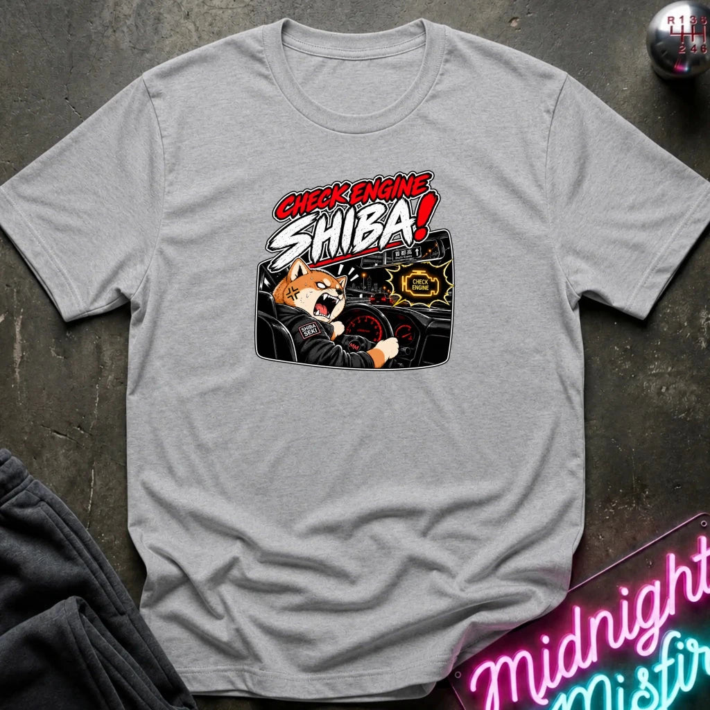 A heather gray short-sleeve t-shirt laid flat on a dark textured surface, featuring a graphic of a Shiba Inu dog driving a car with the text 'CHECK ENGINE SHIBA!' in red and white.