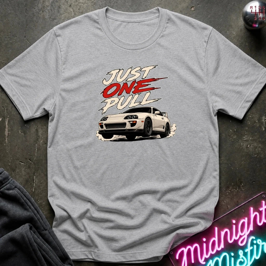 A light gray heather crewneck t-shirt featuring a retro-style graphic. The shirt is displayed flat on a dark textured surface with a neon sign and dark pants partially visible as props.