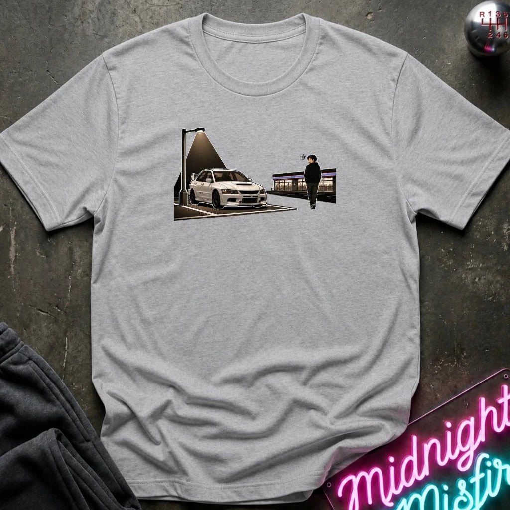 A light gray heather crewneck t-shirt laid flat on a dark textured surface. The shirt features a graphic print in the center showing a white sports car under a streetlamp with a figure standing nearby.