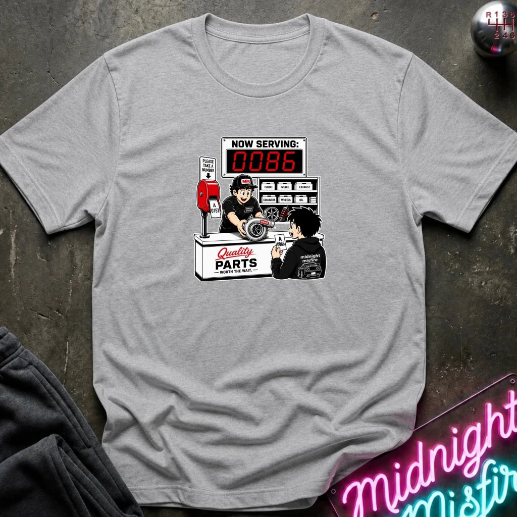 A light gray crewneck t-shirt with a vintage-style graphic of a mechanic working at a 'Quality Parts' counter, displayed on a dark textured background with a neon sign and dark sweatpants.