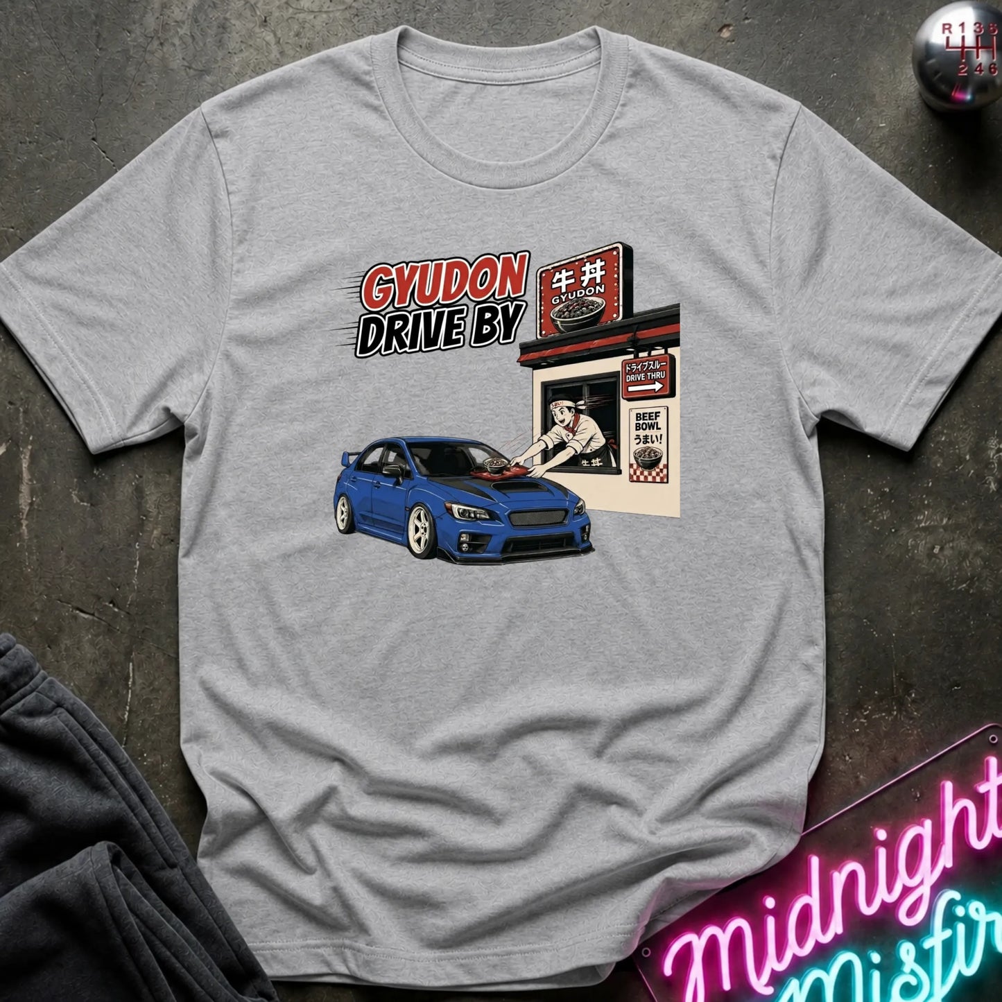 A heather gray crewneck t-shirt featuring a graphic print of a blue sports car and a Japanese-style garage with the text 'GYUDON DRIVE BY'. The shirt is displayed flat on a dark textured surface with a neon sign and dark pants partially visible.