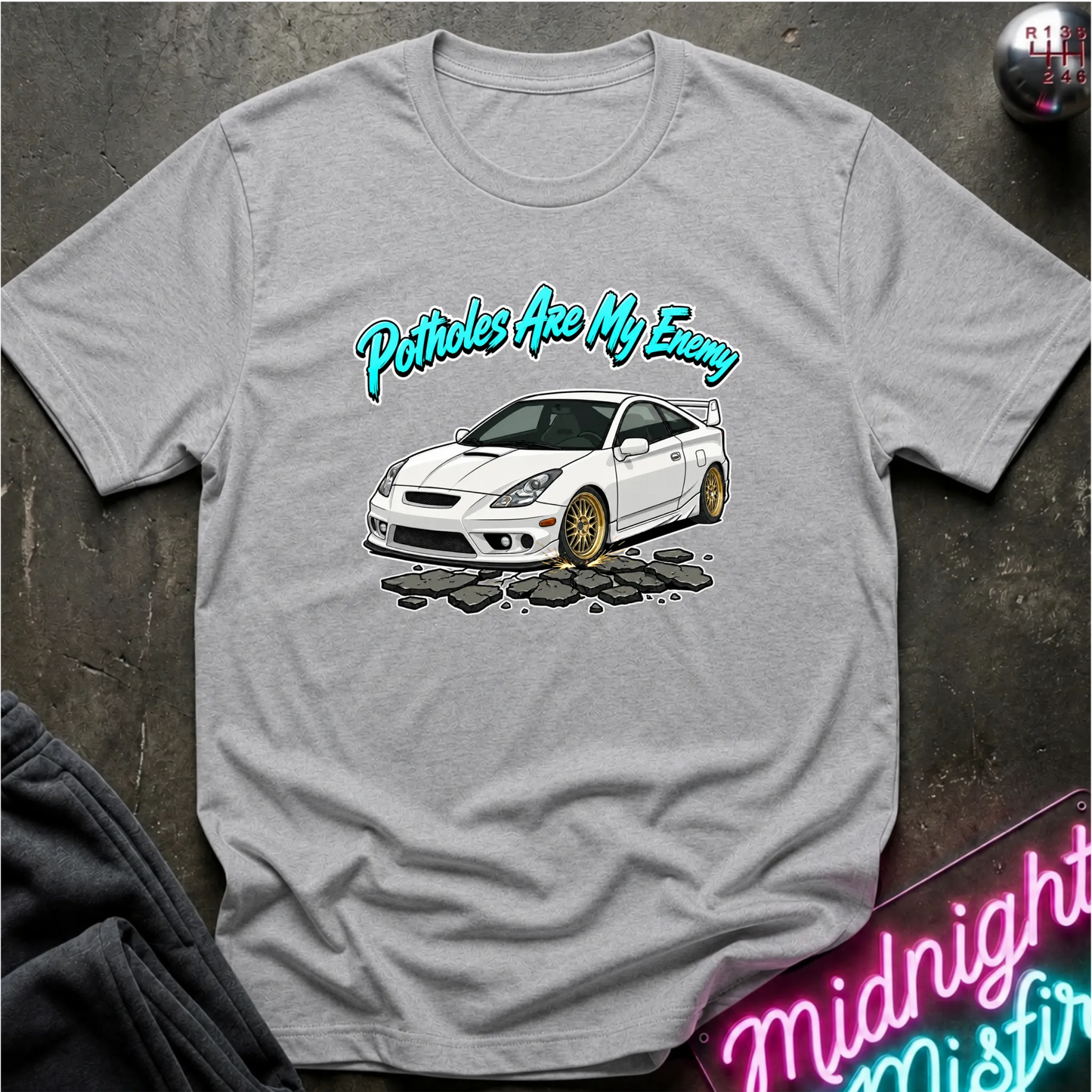 A gray short-sleeve graphic T-shirt laid flat on a dark textured surface. The shirt features a white car illustration with gold wheels and the phrase 'Potatoes Are My Enemy' in teal script above it.