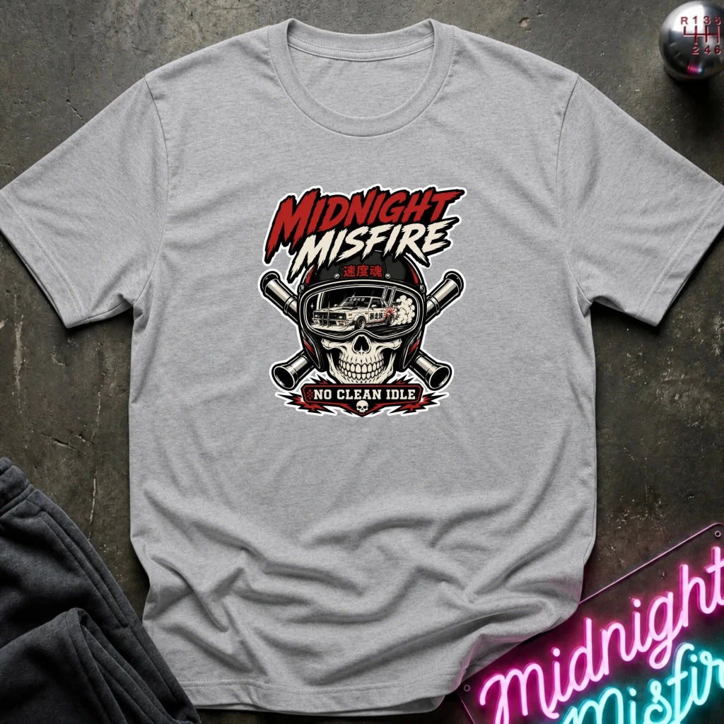 A light gray heather graphic t-shirt laid flat on a dark textured surface. The shirt features a central graphic of a skull wearing a racing helmet with crossed pipes behind it, and the text 'MIDNIGHT MISFIRE' and 'NO CLEAN IDLE'.