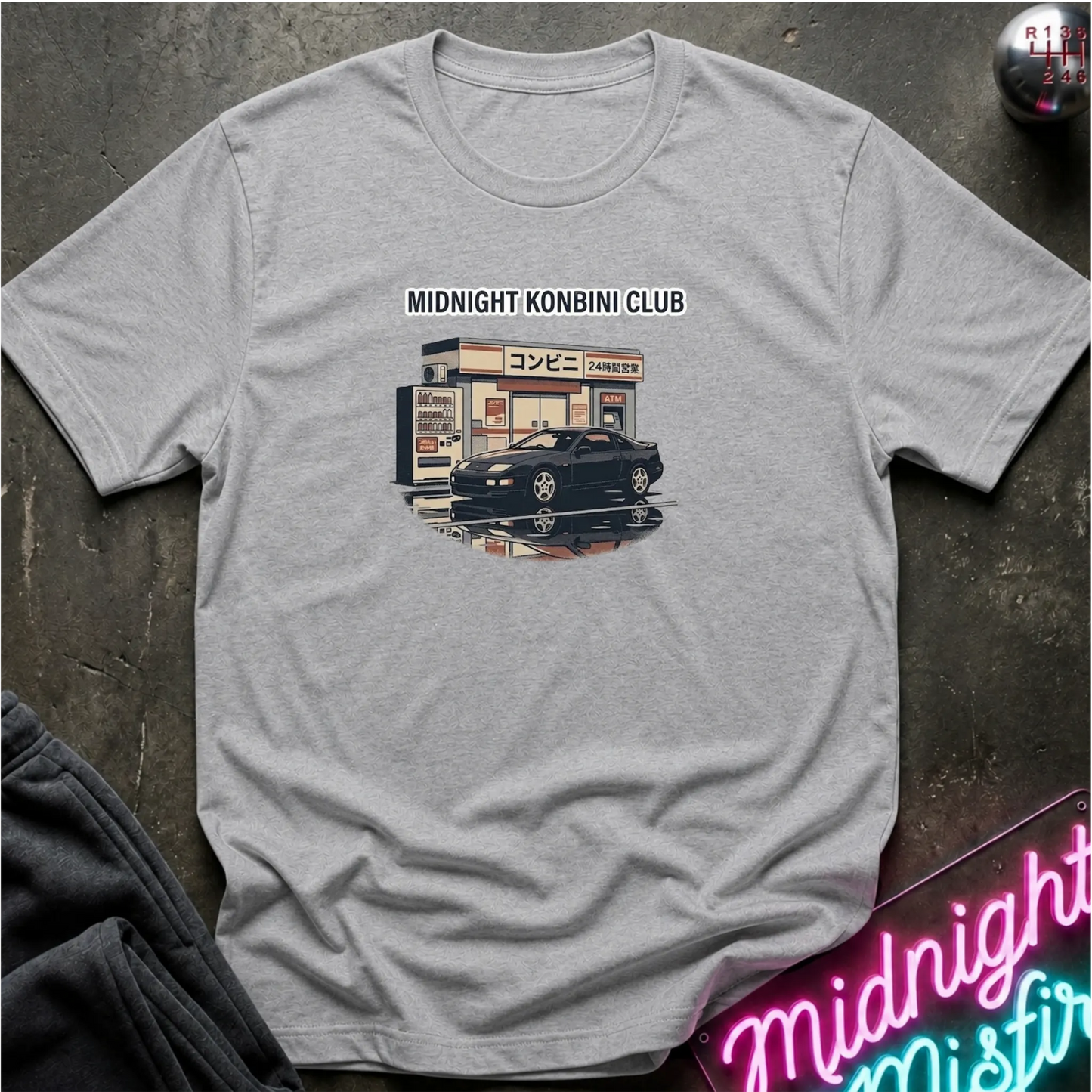 A light gray crewneck t-shirt laid flat on a dark textured surface, featuring a graphic print of a Japanese convenience store with the text 'MIDNIGHT KONBINI CLUB' above it. A black sports car is parked in front of the store.