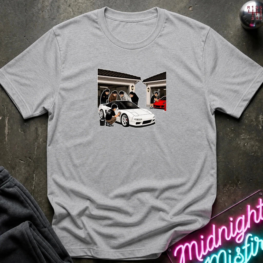 A light gray heather crewneck t-shirt featuring a rectangular graphic print of a white sports car and people, laid flat on a dark textured surface with a neon sign and dark sweatpants partially visible.
