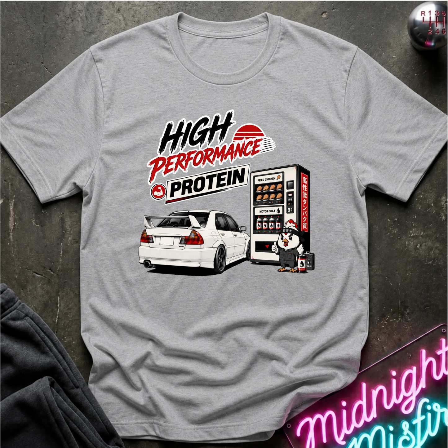 A heather gray short-sleeve t-shirt with a graphic print featuring the text 'HIGH PERFORMANCE PROTEIN' and an illustration of a white sports car next to a vending machine. The shirt is laid flat on a dark textured surface.