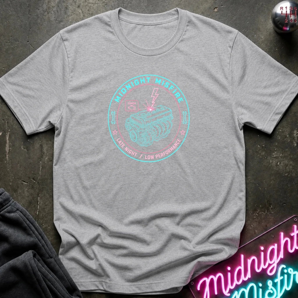 A heather gray short-sleeve t-shirt with a circular graphic on the chest reading 'Midnight Misdre Late Night / Low Performance' in pink and teal, styled flat on a dark textured surface with a neon sign and dark pants partially visible.