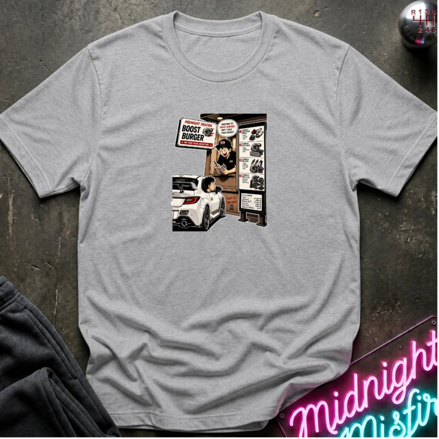 A heather gray short-sleeve crewneck t-shirt laid flat on a dark textured surface, featuring a central graphic print of a street-food burger scene with a white car and signs. 