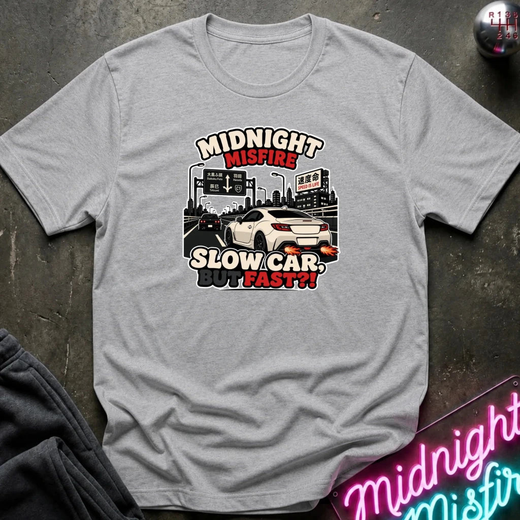 A light gray crewneck t-shirt with a graphic print of a white sports car on a highway and the text 'MIDNIGHT MISFIRE SLOW CAR BUT FAST!!' in red and black lettering. The shirt is displayed flat on a dark textured surface with a neon sign and dark pants partially visible.