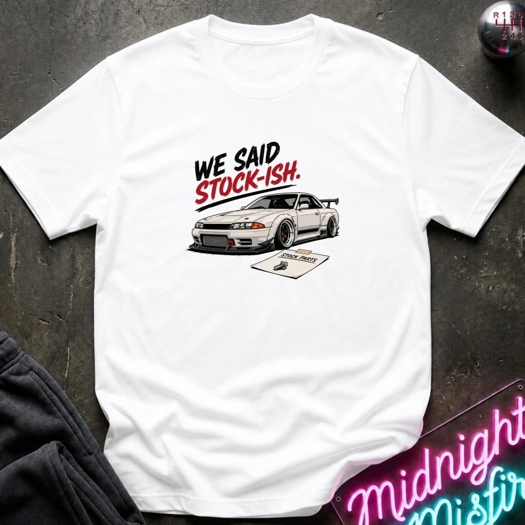 A white short-sleeve graphic T-shirt laid flat on a dark textured surface. The shirt features a printed design of a white sports car with the text 'WE SAID STOCK-ISH.