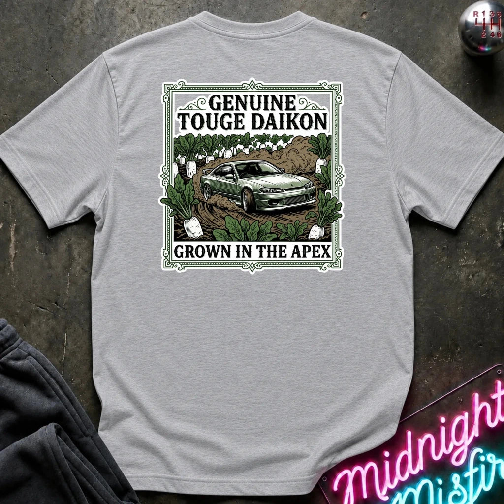 A light gray short-sleeve graphic T-shirt laid flat on a dark textured surface. The shirt features a rectangular vintage-style print with the words 'Genuine Touge Daikon' and 'Grown in the Apex' surrounding an illustration of a green car in a field.