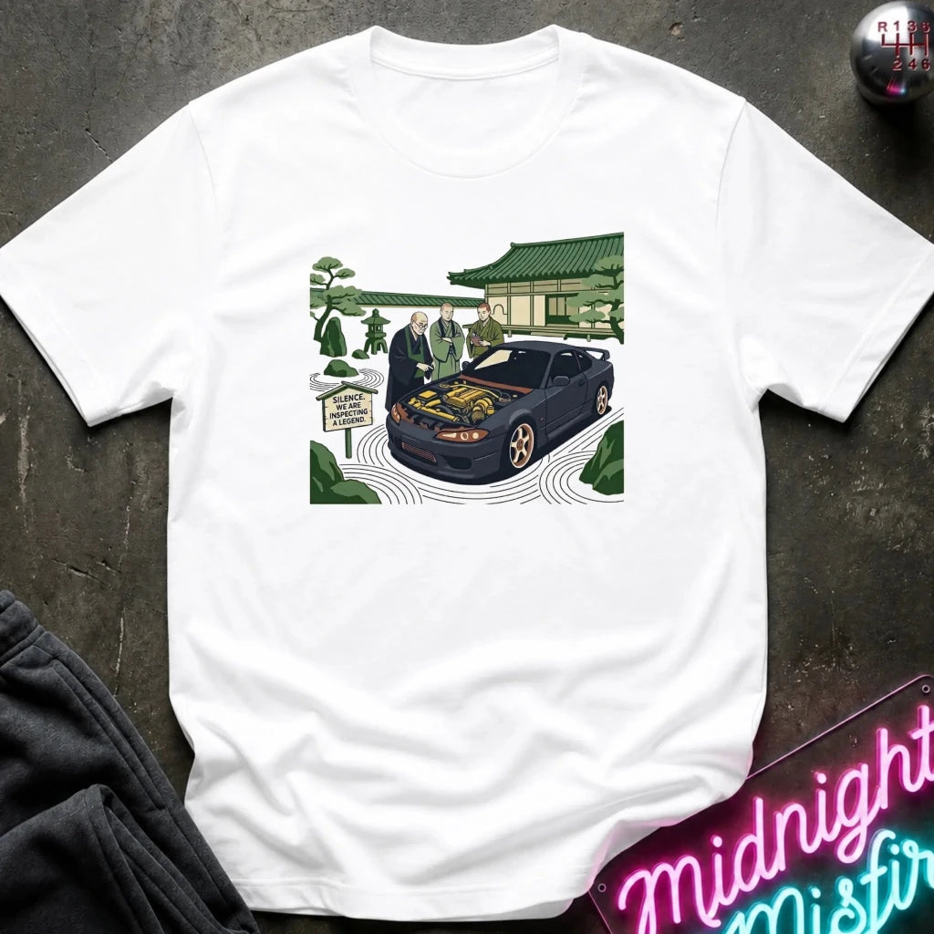 A white short-sleeve crewneck t-shirt featuring a colorful graphic print of a black sports car with its hood open, surrounded by people in a garden setting. The shirt is displayed flat on a dark textured surface with a neon sign and dark pants partially visible.