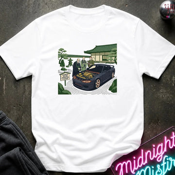 A white short-sleeve crewneck t-shirt featuring a colorful graphic print of a black sports car with its hood open, surrounded by people in a garden setting. The shirt is displayed flat on a dark textured surface with a neon sign and dark pants partially visible.