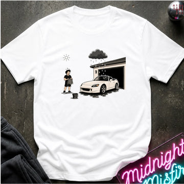 A white short-sleeve graphic T-shirt laid flat on a dark textured surface. The shirt features a printed illustration of a person standing next to a white convertible car in front of an open garage, with a rain cloud above and a sun icon to the left.