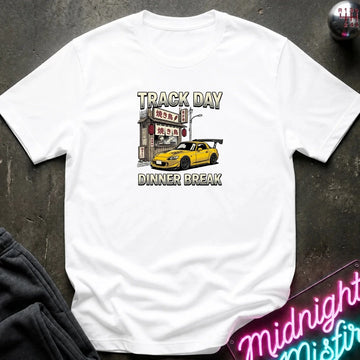 A white short-sleeve graphic t-shirt laid flat on a dark textured surface. The graphic features a yellow sports car parked in front of a Japanese-style restaurant with the text 'TRACK DAY DINNER BREAK' written in a distressed font.