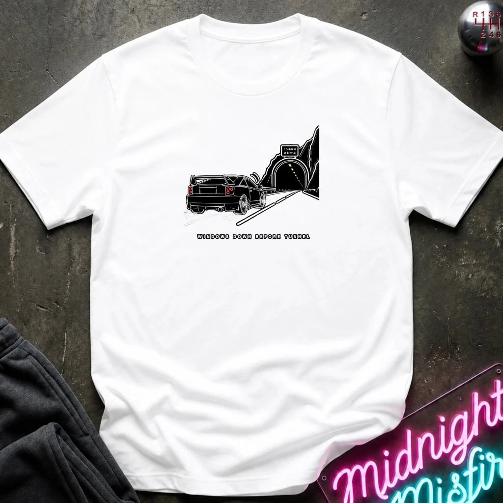 A white short-sleeve t-shirt laid flat on a dark textured surface, featuring a black graphic of a classic car in a tunnel with the text.