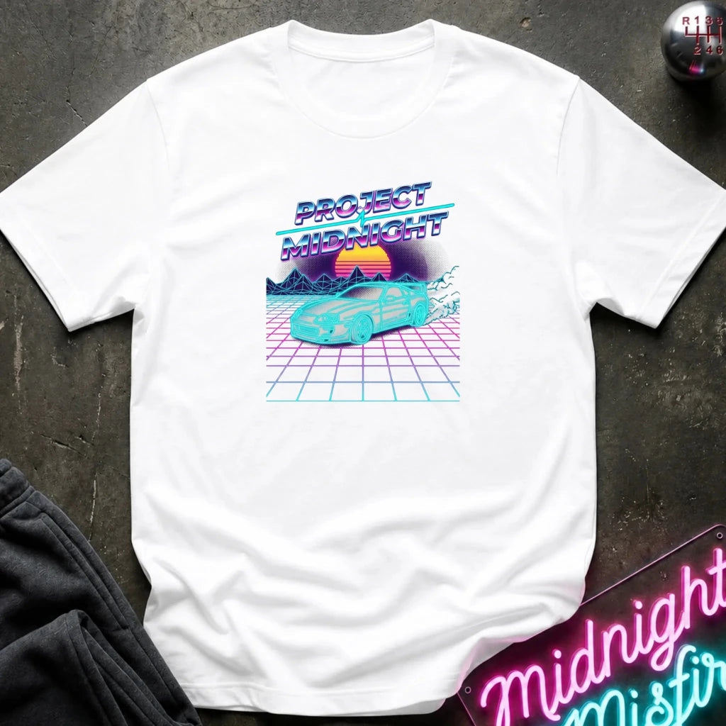 A white short-sleeve t-shirt with a retro 80s synthwave graphic reading 'PROJECT MIDNIGHT' above a turquoise car on a pink grid with a sunset, laid flat on a dark textured surface with a neon sign and dark pants partially visible.