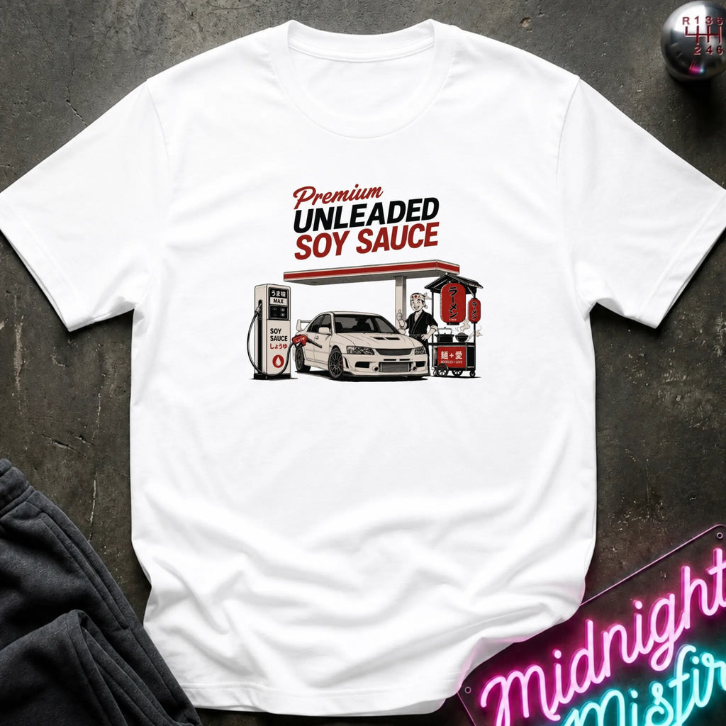 A white short-sleeve graphic t-shirt laid flat on a dark textured surface. The graphic features a vintage-style illustration of a gas station attendant filling up a white car with soy sauce, with the text 'Premium UNLEADED SOY SAUCE' above it.