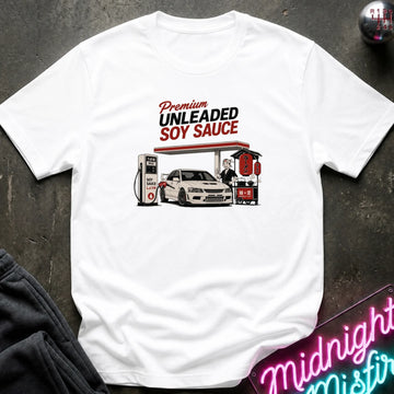 A white short-sleeve graphic t-shirt laid flat on a dark textured surface. The graphic features a vintage-style illustration of a gas station attendant filling up a white car with soy sauce, with the text 'Premium UNLEADED SOY SAUCE' above it.