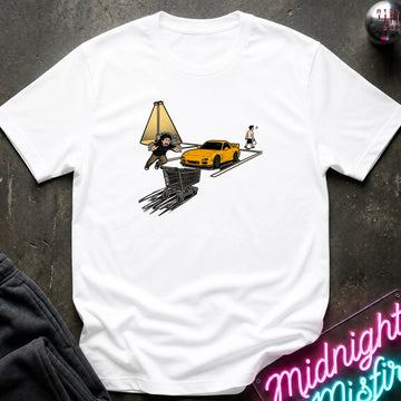 A white short-sleeve graphic t-shirt laid flat on a dark textured surface. The graphic shows a yellow sports car, a shopping cart, a streetlamp, and two people in a surreal, sketch-like illustration.